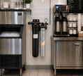 Commercial kitchen equipment setup with a stainless steel ice machine on the left, a wall-mounted water filter system in the center, and a coffee station with two coffee makers and white mugs on the right.